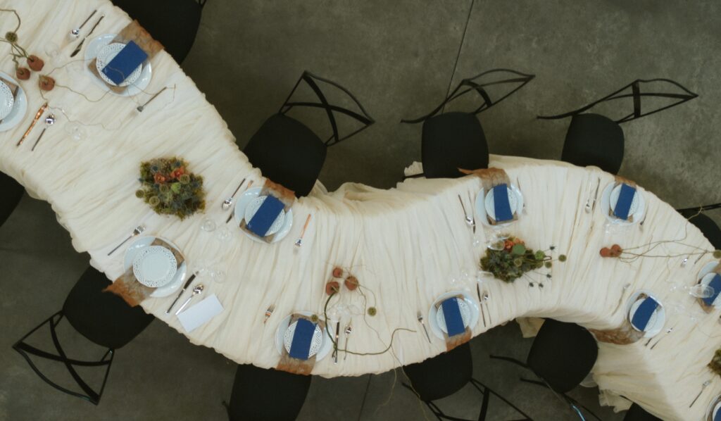 Detailed overhead perspective of a curved wedding table setup with organic florals and modern black x-back chairs.