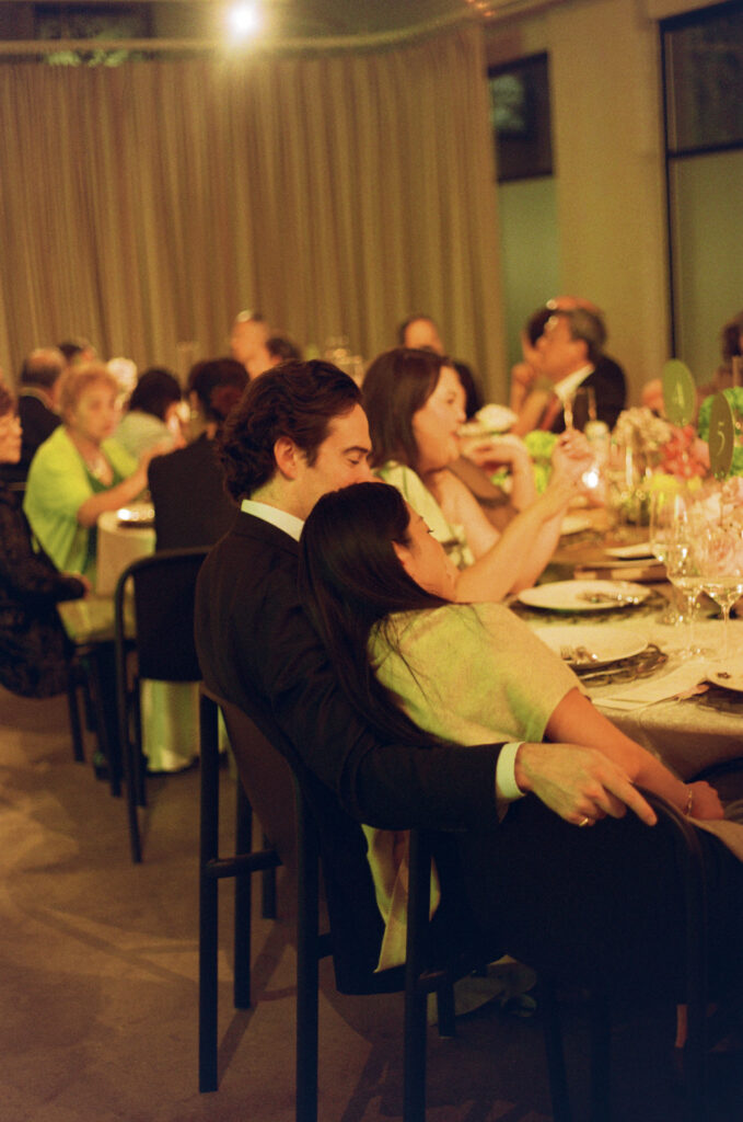 Bride and groom embracing at a candlelit wedding reception table with modern black chairs and lush pink peony centerpieces.