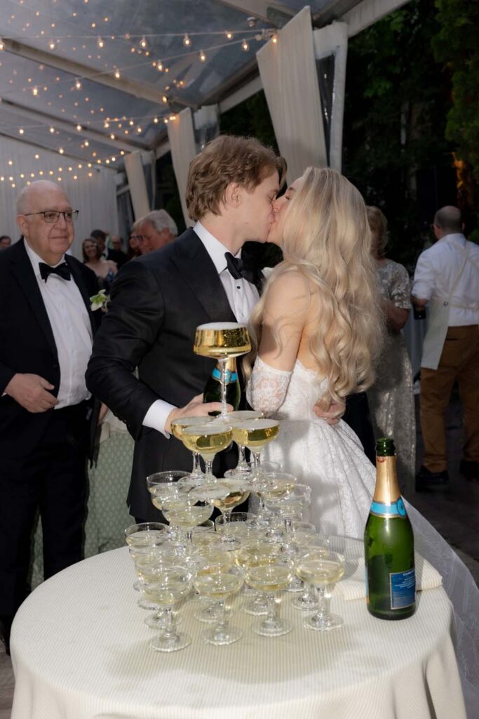 Bride and groom kissing behind a completed champagne tower at an outdoor wedding event.