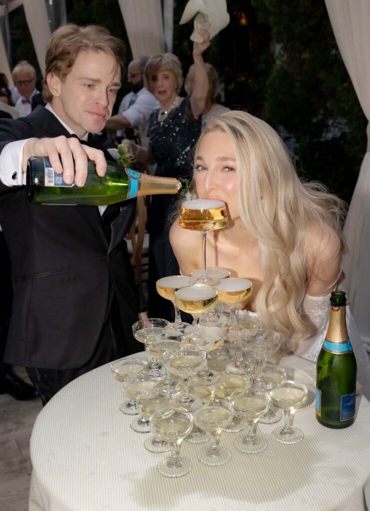 Bride drinking from the top coupe of a champagne tower after pouring.