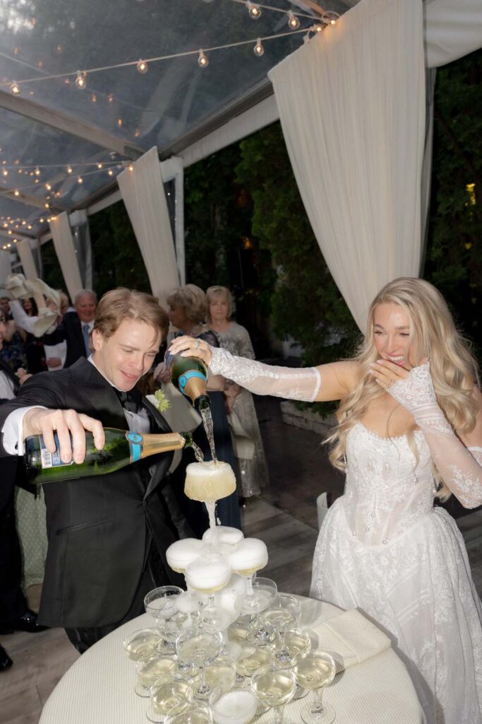 Bride and groom pouring champagne onto the top of a champagne tower at a wedding reception.