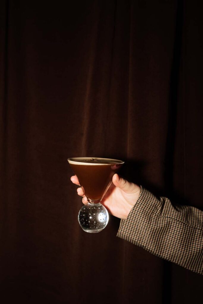 Hand holding an espresso martini in a clear ball martini glass against a dark brown velvet backdrop.