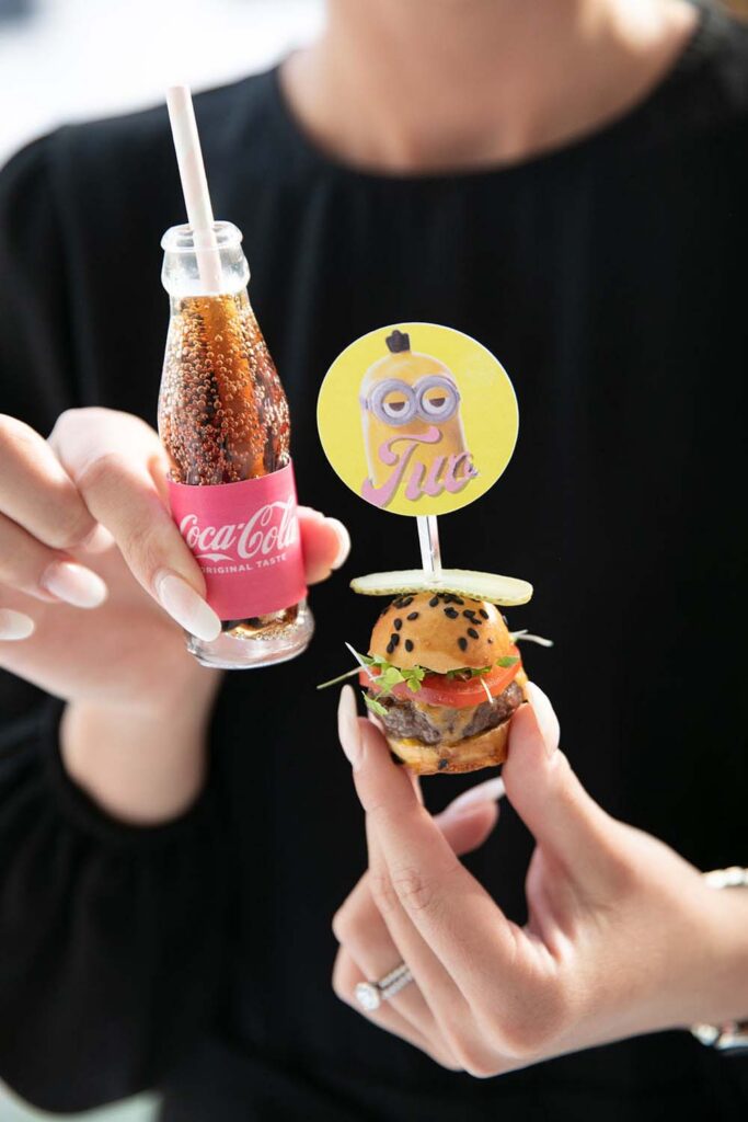 Guest holding a mini Coca-Cola bottle alongside a mini slider hors d'oeuvre.