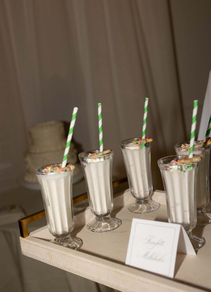 Mini milkshakes served in mini parfait glasses with green striped straws on a leather tray.