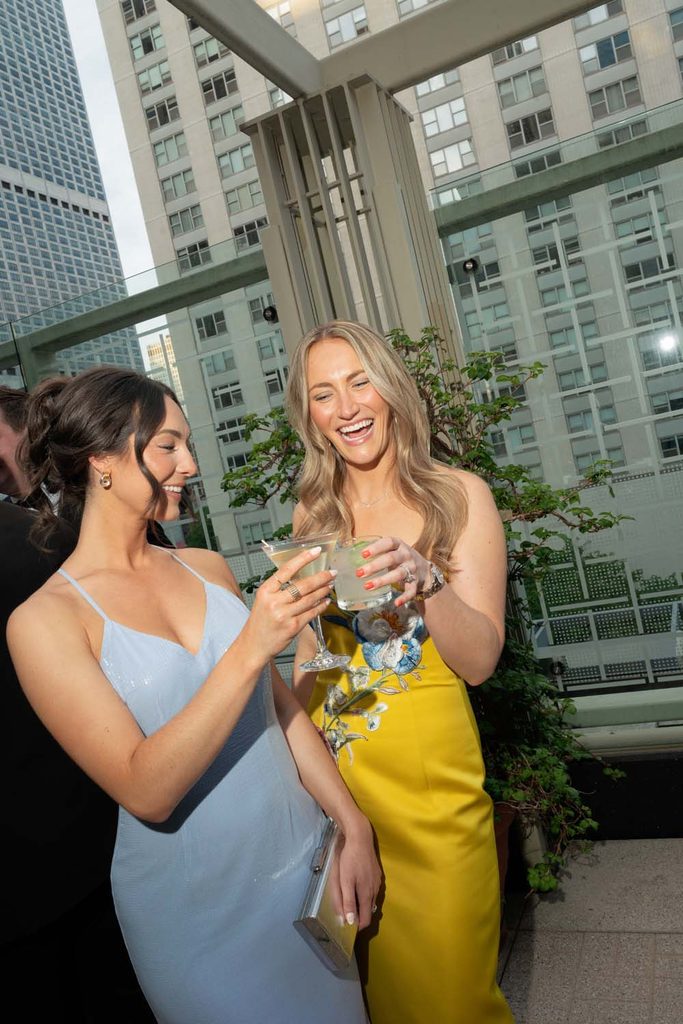Two smiling event guests laughing and toasting with cocktails on an outdoor rooftop terrace with Chicago high-rises in the background