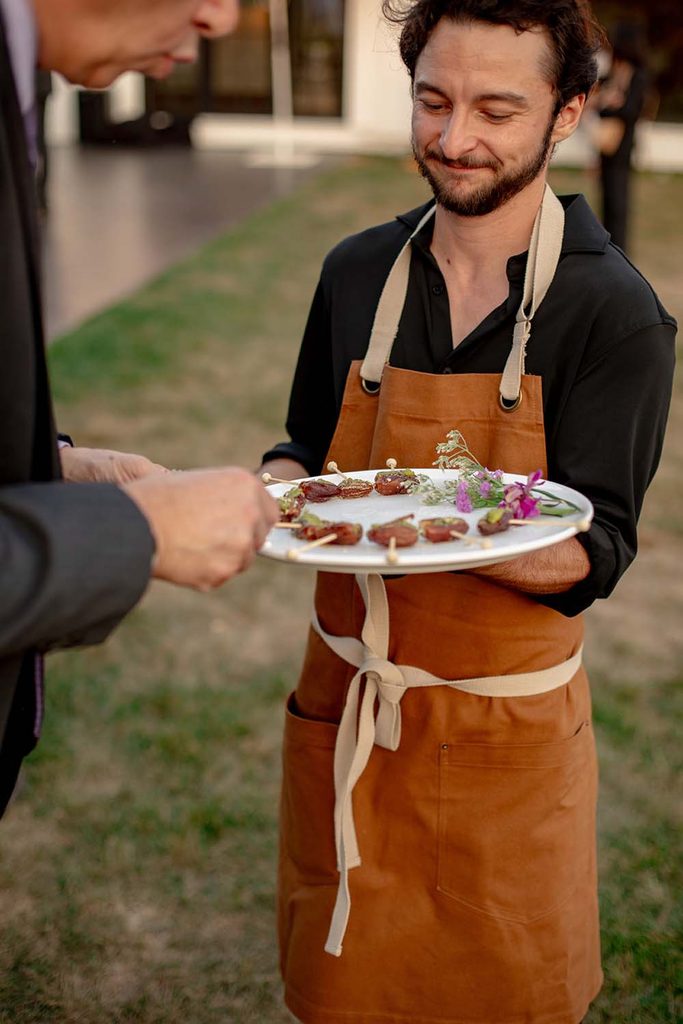 Server in a brown apron offering a round platter of skewered appetizers garnished with fresh flowers to a guest.