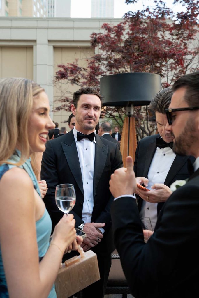 Guests in formal evening wear and black-tie tuxedos socializing at an outdoor rooftop cocktail hour.