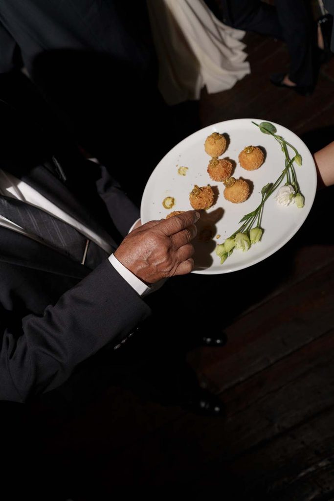 Overhead view of a guest selecting a fried arancini appetizer from a butler-passed white serving platter garnished with white flowers.