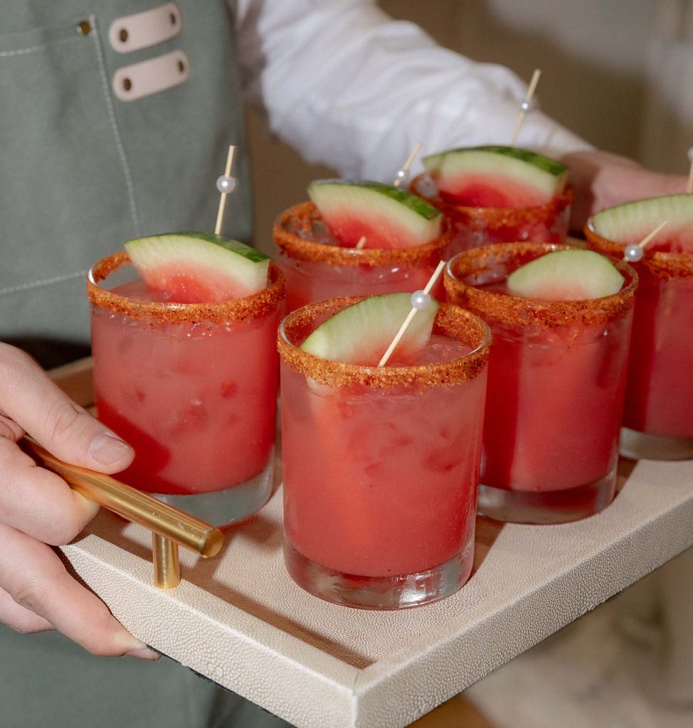Close-up of a server carrying a tray of watermelon cocktails with chili-rimmed glasses and watermelon wedge garnishes.