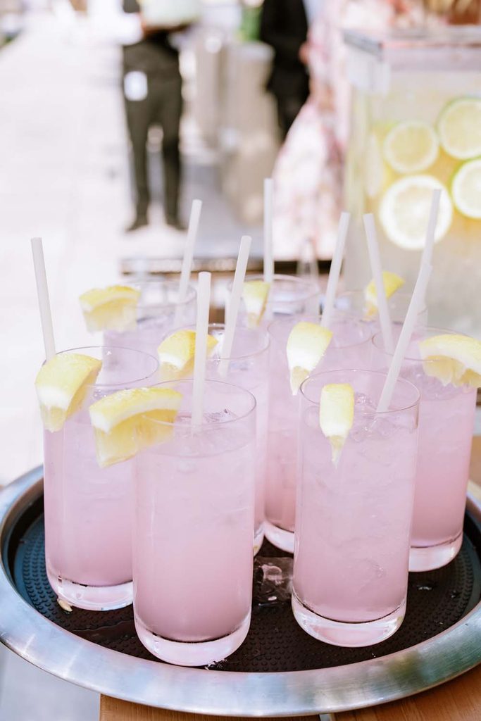 ray filled with tall glasses of iced pink lemonade garnished with fresh lemon wedges and straws at a catered event.